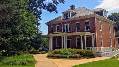 Large two story brick house with a large porch and greenery in front.