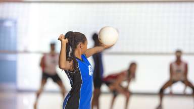 A teenaged girl in a blue jersey is playing in a volleyball match. She is about the serve the ball. They are playing in a school gym.