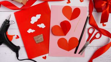 Two hand made valentine cards with red hearts glued on pink construction paper.  Next to the cards are a glue gun, sissors and ribbons.