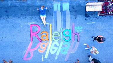 An overhead view of chalk artist Taylor sitting near her chalk art that reads Raleigh with each letter being a different color of the rainbow