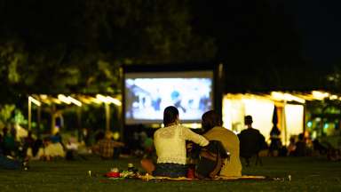 A couple sitting on a blanket watching a movie in a park at night.