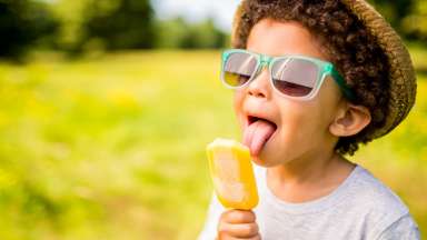 A young boy wearing a straw hat and sunglasses licking a popsicle.