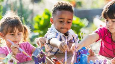 a group of preschoolers sit at a table outside and paint