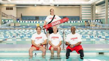 Four Lifeguards posing for a picture sitting on the edge of a pool.