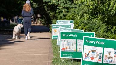 Person walking a dog with story walk signs in the grass beside the sidewalk