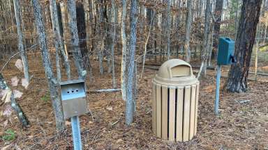 A park-like setting with a garbage can, pet waste bag dispenser, and cigarette butt disposal box