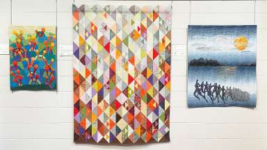 Three quilts by members of the Professional Art Quilters Alliance - South hang on a white wall at Sertoma Arts Center