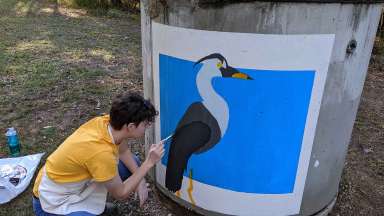 An artist paints a bird on a sewer up next to a Raleigh greenway