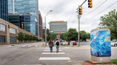 Two people walk on a crosswalk in downtown Raleigh next to a signal box vinyl wrapped with artwork by Sam Greene