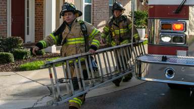 Two Raleigh firefighters carry a ladder in front of a residential home