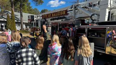 School group listens to firefighters talk as they show children a ladder truck.