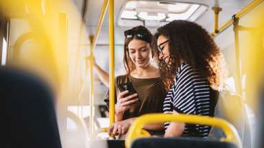 Two teenage girls standing while riding a bus. One girl looking at her phone