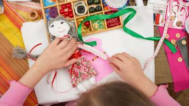 an image of a girl measuring a dolls arm with buttons, beads, yarn, ribbon and other crafts on a table.