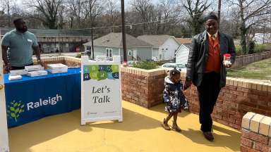 Raleigh employee standing at a table next to a &quot;Let's Talk&quot; sign. A father holding daughter's hand is nearby.