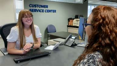 A woman at a desk talking to another person. A Customer Service Center sign in the background