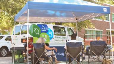 Three people sit in lawn chairs under a ten in front of the Raleigh Engagement Van