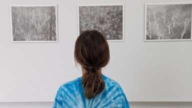 Young white girl with a long pony tail and blue tie dye shirt looking art three pencil drawings on a gallery wall.