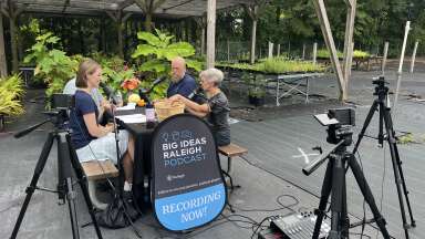 Podcast recording at an urban garden, podcasters are recording at a table with a sign taht says Big Ideas Raleigh Podcast Recording Now in front of them