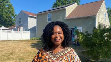 Photo of Wanda Statum standing in her backyard with her home in the background. Blue sky is visible.