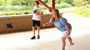 a person tossing a cornhole beanbag while a friend watches