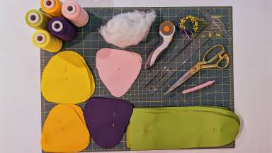 A cutting board covered with sewing tools, fabric and thread, the starting bits of a soft botanical sculpture,