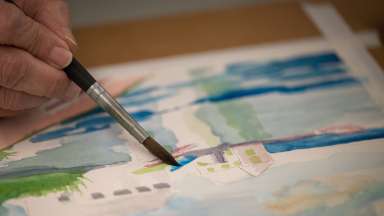 A close up photograph of a hand holding a brush against an unfinished watercolor painting.
