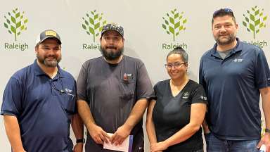 Four City of Raleigh employees stand in front of a board with the Raleigh logo repeating on it.