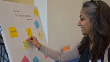 Woman smiling and placing a sticky note on a board that says "What does entrepreneurship mean to you?"