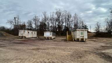 Three dumpsters sitting in dirt area with trees behind.