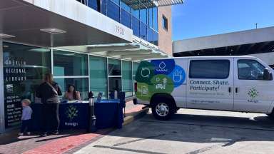 Engagement Van is parked in front of a Wake County Library location. A mother and her child speak to the City staff member at a table beside the van.