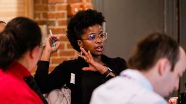 A young black women wearing a black shirt and round glasses participating in a dicussion around a table.