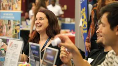 Woman sits behind exhibit table with two of her colleagues, smiling as she talks to a passerby.