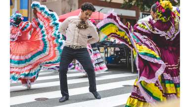 Three dancers in vibrant traditional Mexican attire perform on a crosswalk during Fiesta del Pueblo.