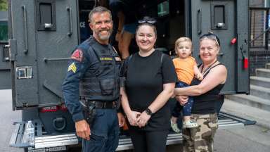 A Raleigh Police officer with two women and a small child standing in front an armored vehiclea smiling at the camera.
