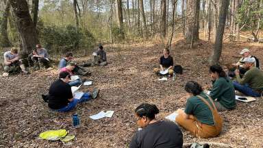 A group of people sits in a circle on the ground in a forest with fall leaves.