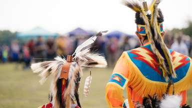 An art photograph of a tall person dressed in Native American headpiece and a small person/child also with a native American headpiece standing next to each other