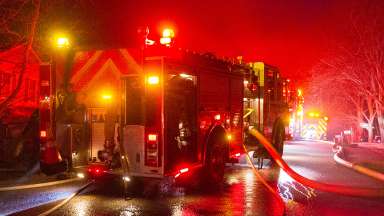 Looking at the back and side of a large fire engine with water hose connected on side of truck at night.