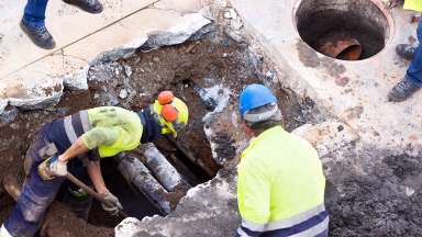 Construction works working in a hole in pavement with pipes visible.