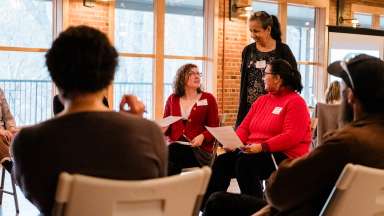 A group of people sitting in chairs with a person standing talking to one of the participants
