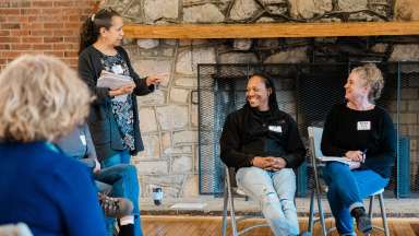 a stone fireplace in backgroun with people in charis and someone standing up talking to them.