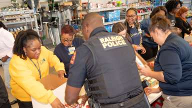 Police officers along with other volunteers stand at a long table packing boxes with food