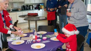 A child dressed in a Santa hat and a red sweater sits at a table decorating a cookie. Adults look on smiling.