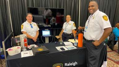 Three Raleigh Fire personnel stand at a exhibit education table with flyers, a tv behind the, and a fire extinguisher on the table.