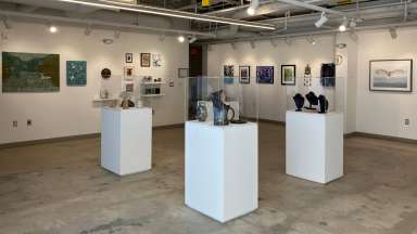 A wide view of a gallery space showing photographs and paintings hanging on walls and pedestals with pottery and jewelry