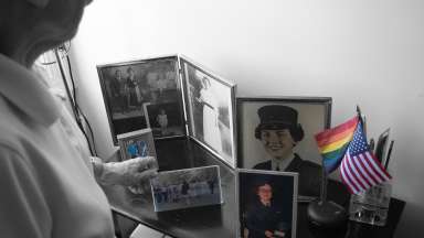 black and white photograph of person looking at family photos displayed on side table, with select portraits in color, alongside American and pride flags in color