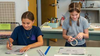 A photograph of two smiling campers working on printmaking projects at a table in the printmaking studio.