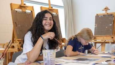 A photograph of two campers painting with acrylics at a table in a studio.
