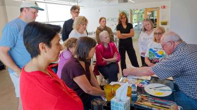 Instructor Rick Bennett, an older white male with glasses and wearing a blue gingham shirt and jeans, is demonstrating a watercolor technique for a group of male and female students of various ages