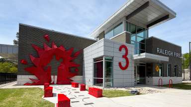A red metal sculpture with the outline of a firefighter rescuing a woman is on the wall of a fire station building.