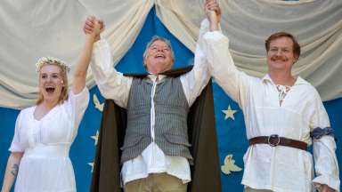 three actors on an outdoor stage joining hands and smiling